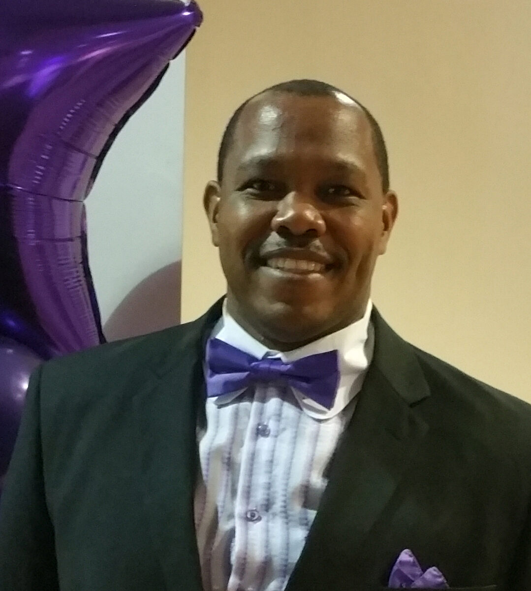 Chris Davios in a dark suit with purple bow tie and pocket square, smiling warmly